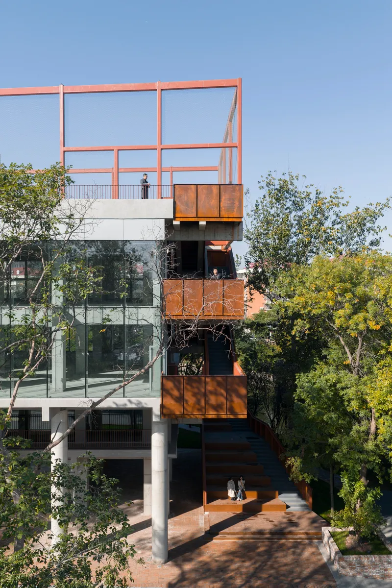 Stacked volumes with weathering steel panels and glass balconies rising through surrounding trees under blue sky