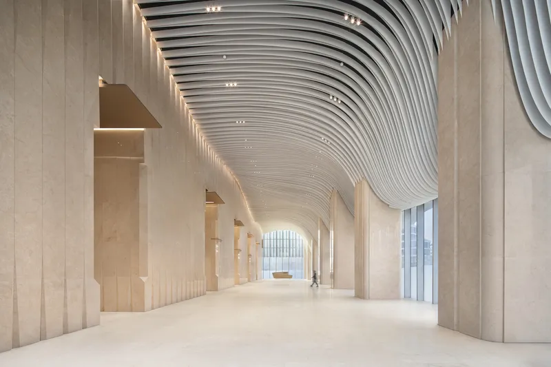 Interior corridor with undulating ribbed ceiling and stone-clad columns illuminated by recessed linear lighting