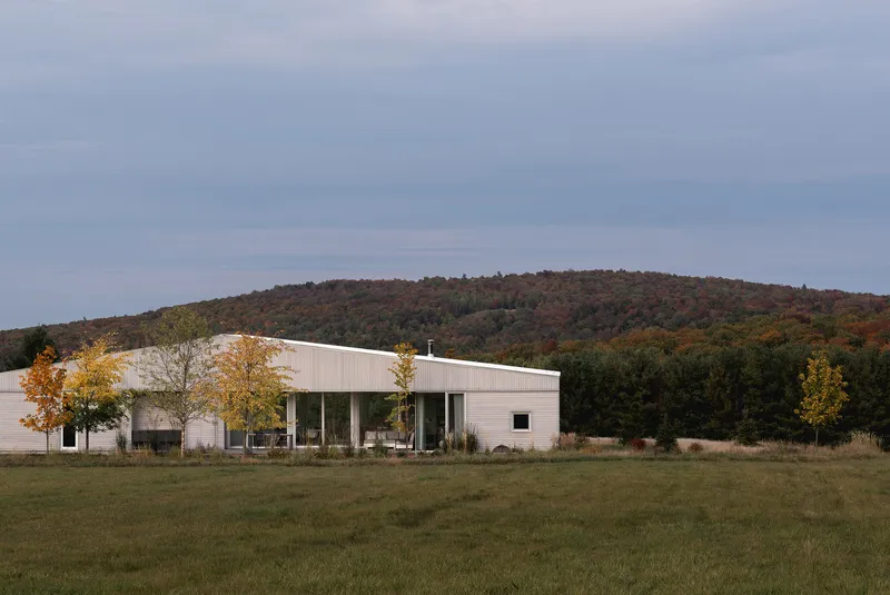 Long view of the white metal-clad residence set in an open meadow with forested hills beyond