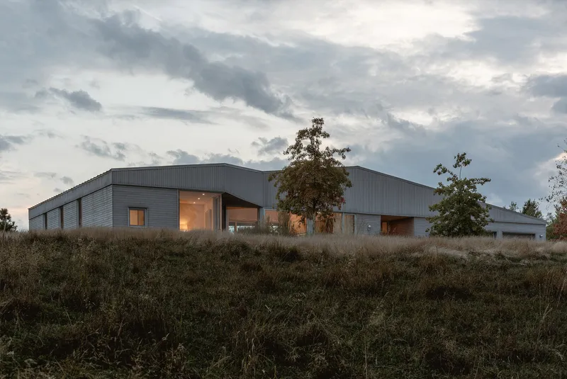 Metal-clad volumes with illuminated glazing at dusk viewed from a sloping meadow with trees