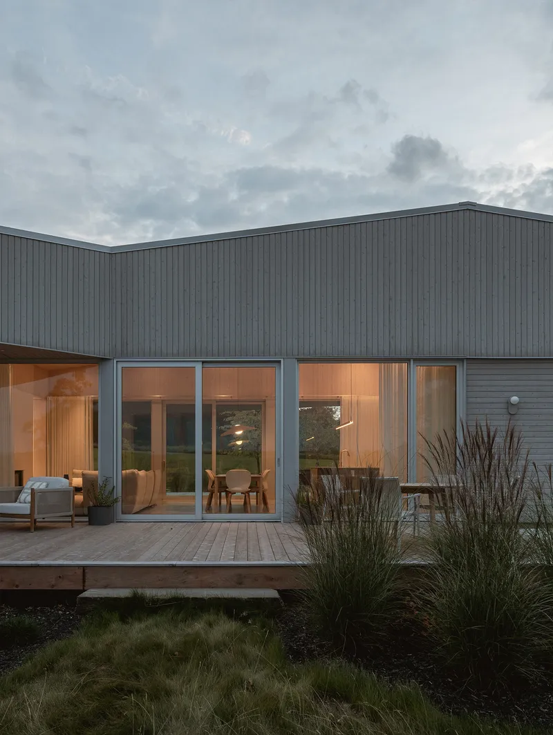 Vertical wood-clad facade with full-height glazing opening onto a timber deck surrounded by native grasses at dusk