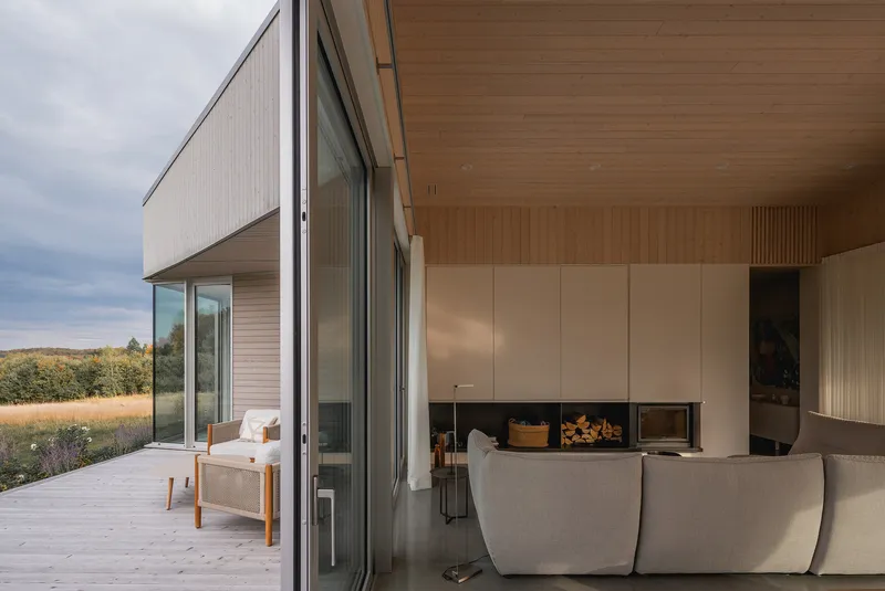 Living area with timber-lined ceiling and pale stone wall housing a fireplace opening to a deck with meadow beyond