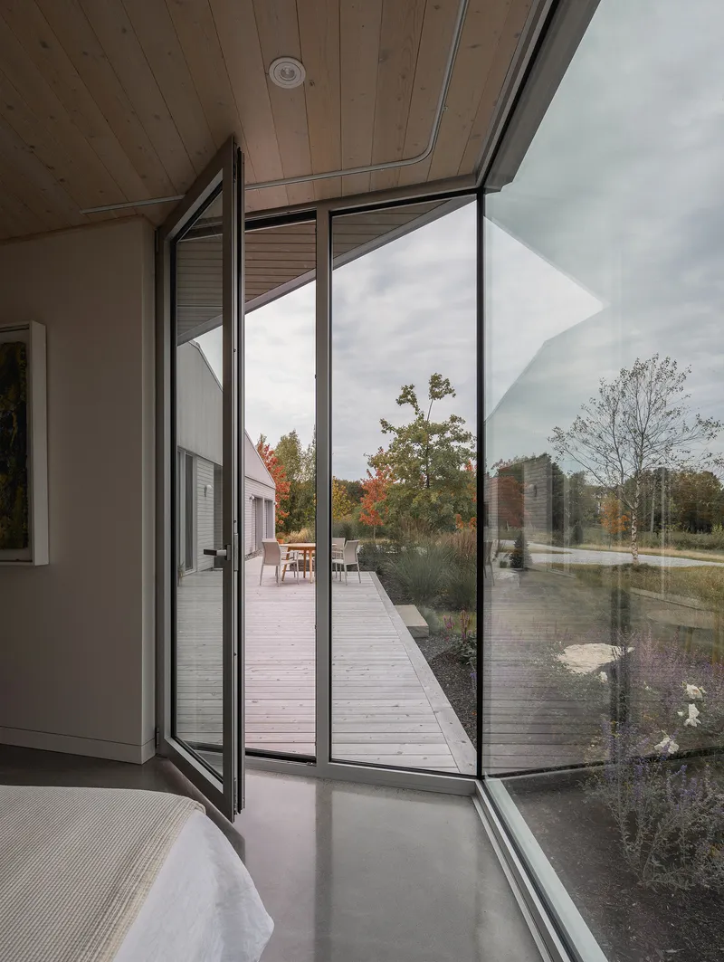 Pivoting glass door opens from bedroom onto timber deck framed by autumn foliage and cloudy sky