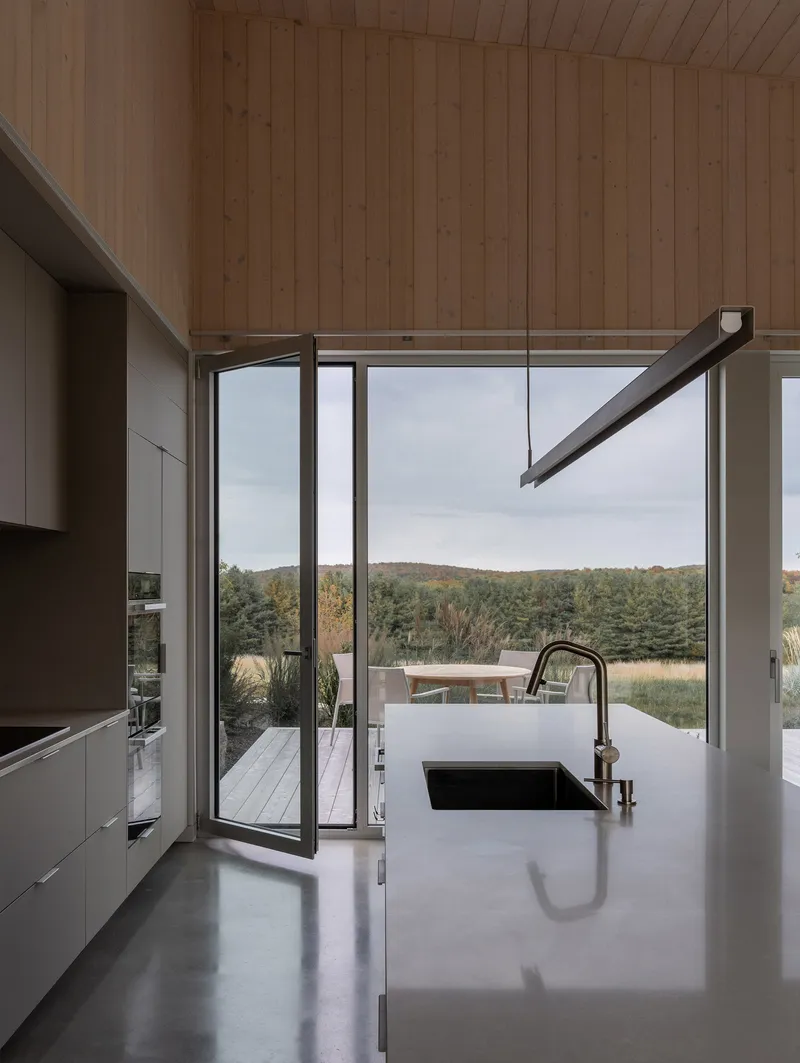Kitchen island with integral sink facing narrow glass door and distant wooded hillside under overcast sky