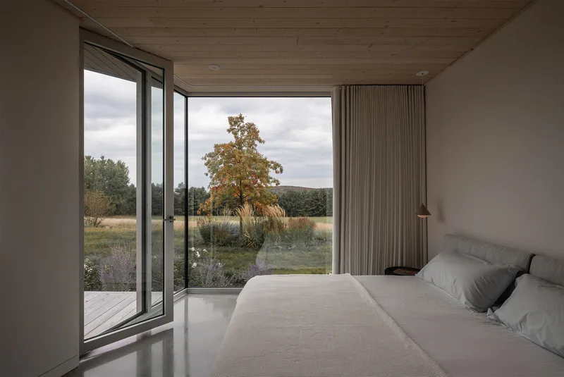 Bedroom with full-height glazing and pleated curtain panel framing a solitary tree in an autumn field