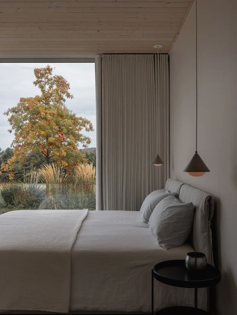 Bedroom corner with pendant lights and pleated curtain beside glazing overlooking grasses and an oak tree