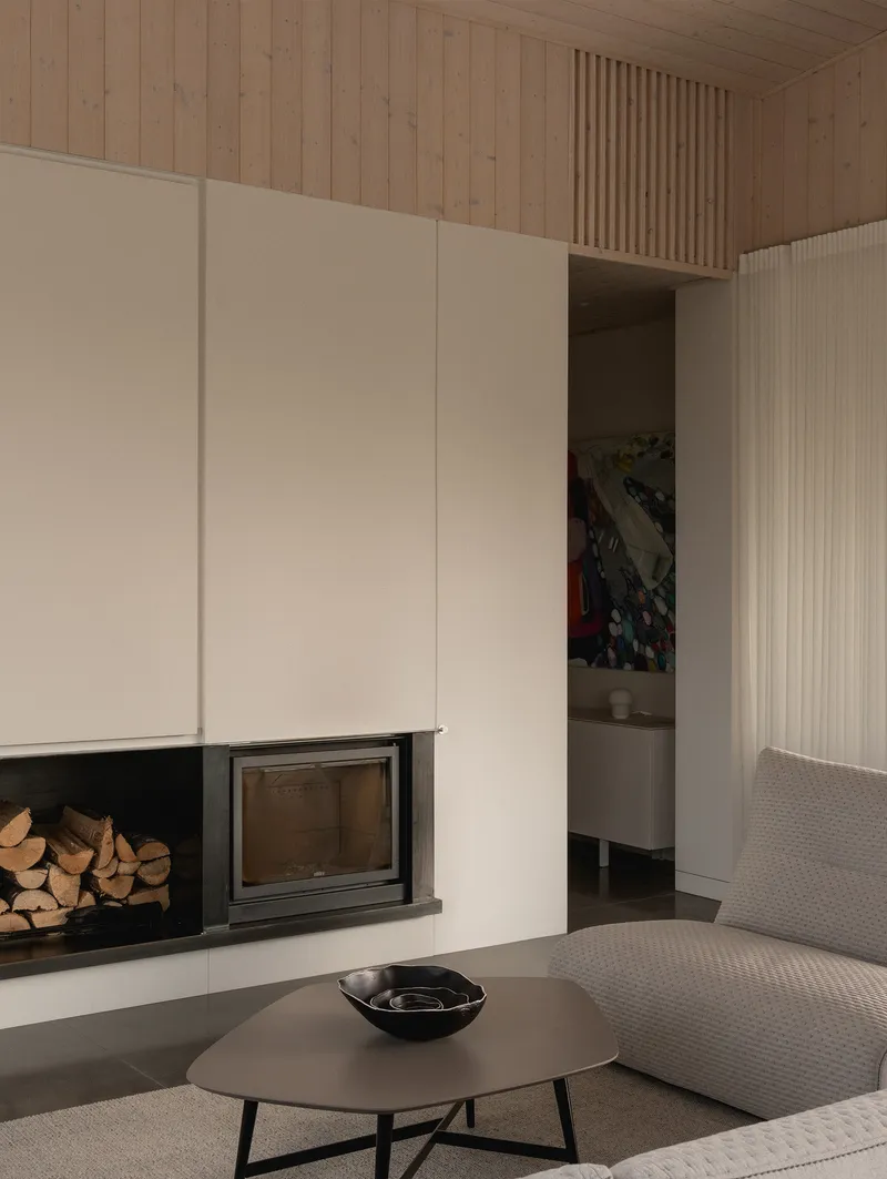 Living room fireplace with stacked firewood storage beneath a timber-lined ceiling volume