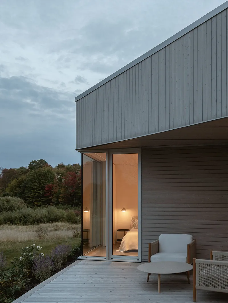 Corner glazing beneath cantilevered upper volume illuminated at dusk overlooking wild grasses and tree line