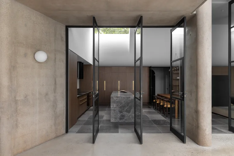 Steel-framed pivot doors opening from concrete corridor into kitchen with stone island and timber millwork