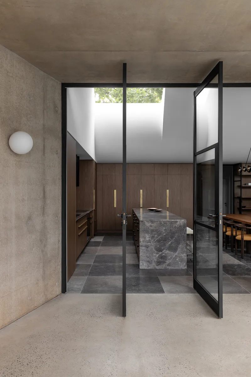 Black-framed pivot door opening to kitchen with dark marble island and clerestory windows above
