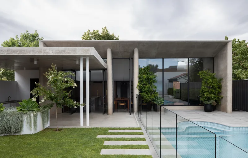 Concrete pavilion with cylindrical columns and full-height glazing overlooking a pool and lawn with stepping stones
