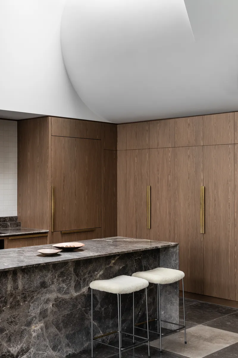 Dark stone island with brass-handled timber cabinetry and upholstered stools in natural light