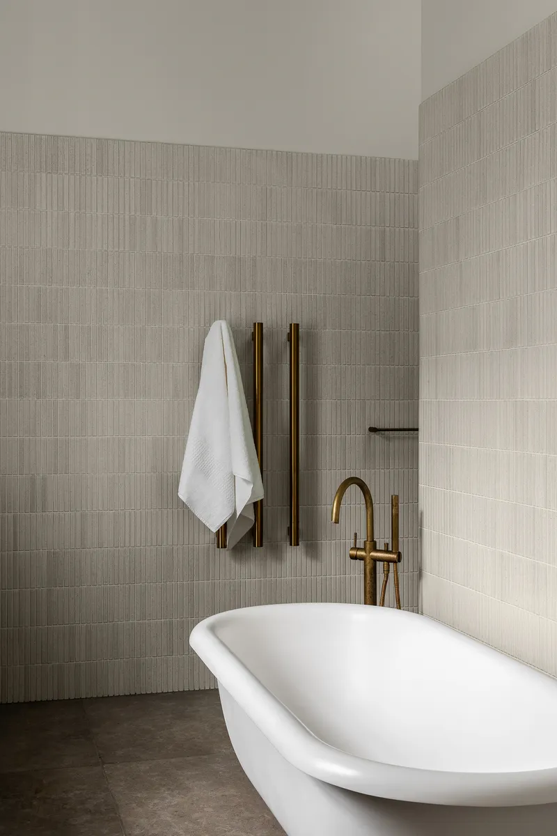 Freestanding white bathtub with brass fixtures against ribbed ceramic tile walls in muted tones