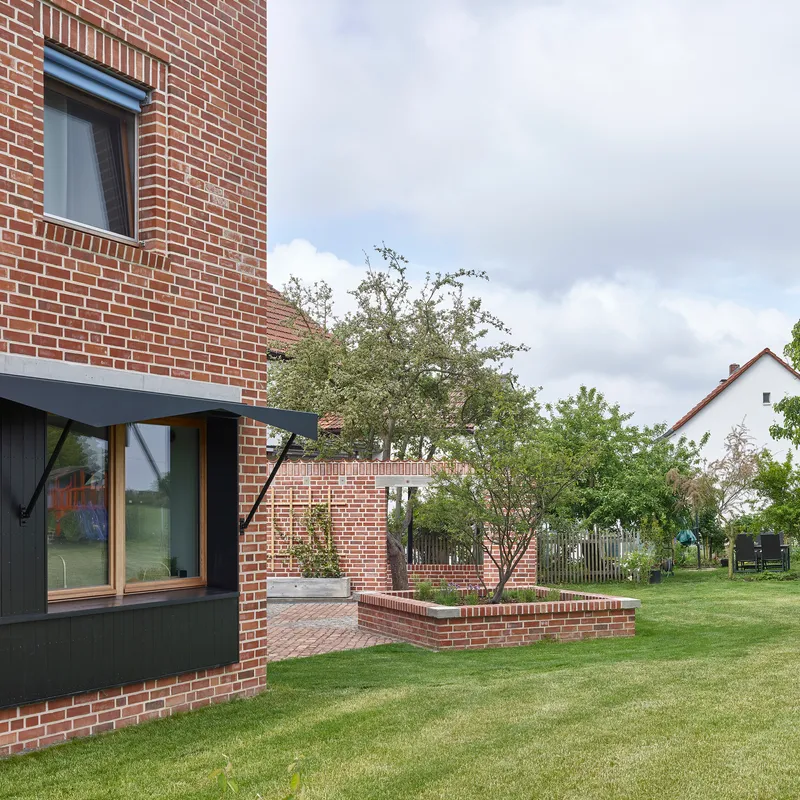 Red brick facade with cantilevered metal awning and planted brick planter beside a lawn with trees