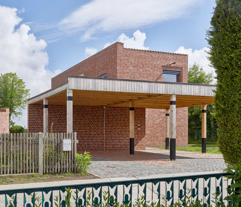Open carport structure with exposed timber beams and columns against brick wall under cloudy sky
