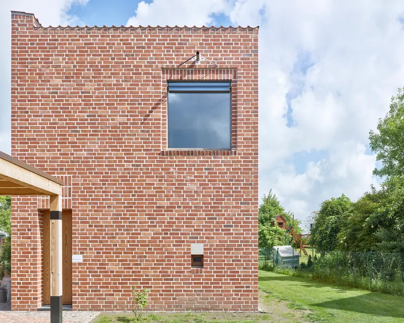 Red brick facade with single recessed window and timber porch entry beneath a cloudy summer sky