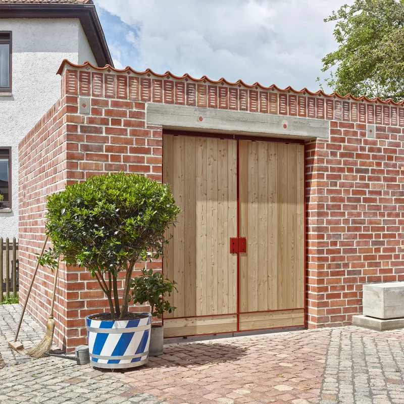 Street facade with double timber doors set below wavy terracotta coping and potted tree in foreground