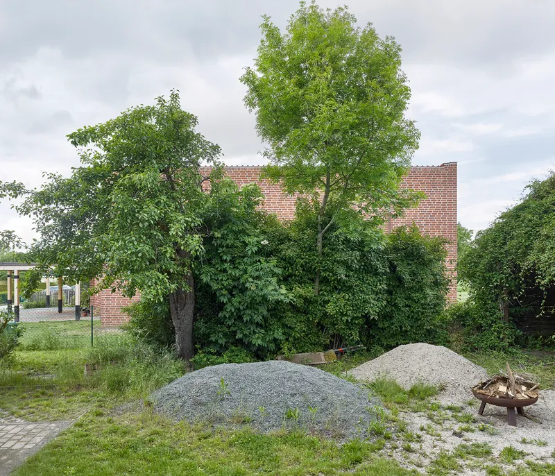 Distant view of the brick volume behind mature deciduous trees and gravel mounds under overcast skies