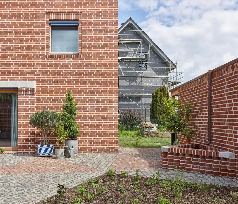 Brick-paved courtyard between red masonry walls with a dark metal gable structure visible beyond under cloudy sky