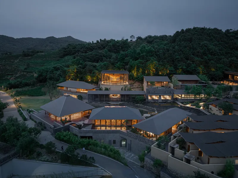 Aerial view of clustered structures with dark pitched roofs nestled into forested hillside at dusk
