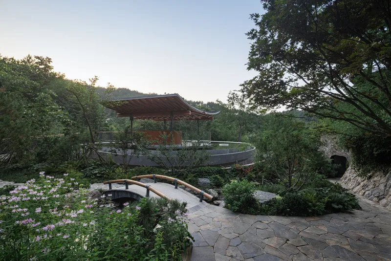 Arched timber footbridge crossing stone-paved garden path toward raised pavilion with broad eaves at dusk