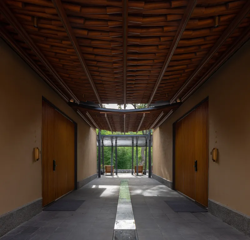 Entry corridor with woven timber ceiling, stone floor with water channel, and green trees visible beyond