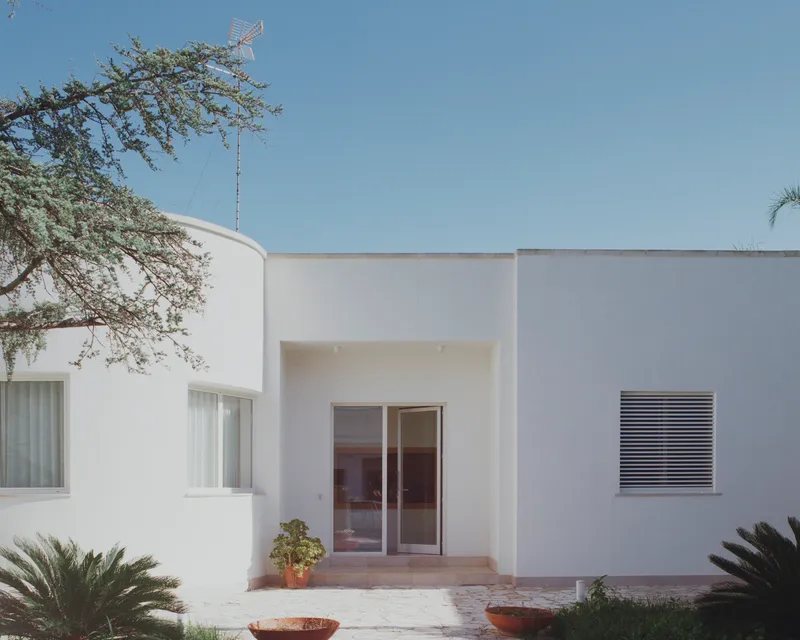 White stucco facade with recessed entry and glazed doors framed by overhanging tree branches and terracotta planters