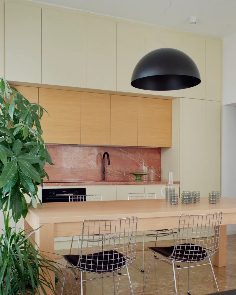 Kitchen with oak and cream cabinetry, pink marble backsplash and wire mesh dining chairs beneath a dome pendant