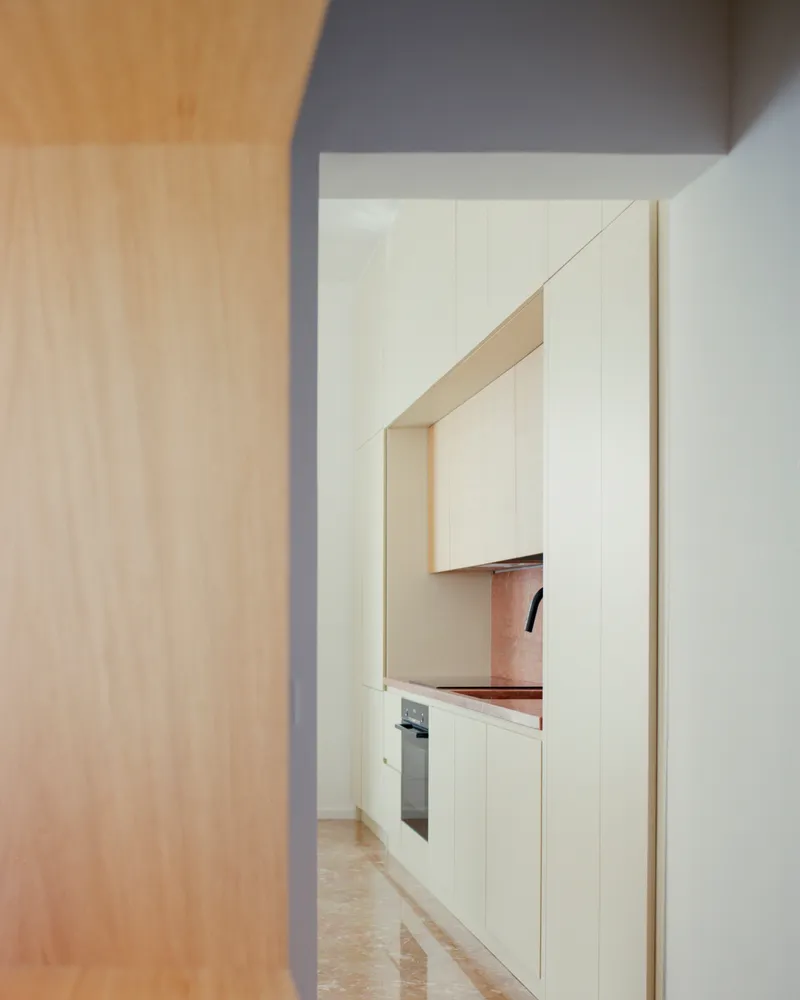 Doorway framed by pale wood paneling revealing kitchen with pink marble countertop and cream upper cabinets