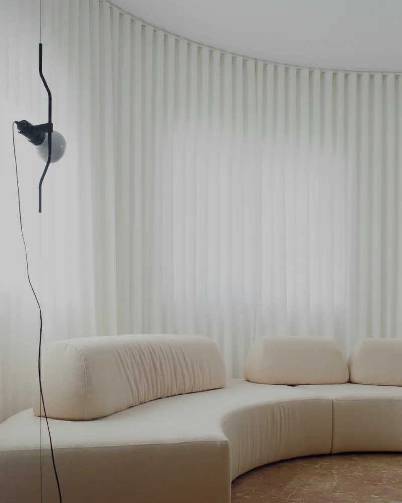 Curved seating upholstered in pale fabric against floor-to-ceiling curtains with a wall-mounted black reading lamp