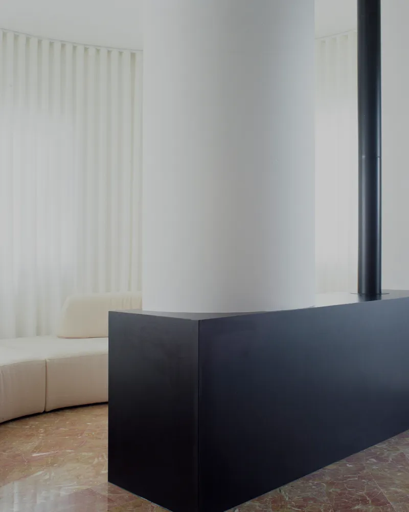 Black steel fireplace hearth next to curved sofa with white curtain and stone floor