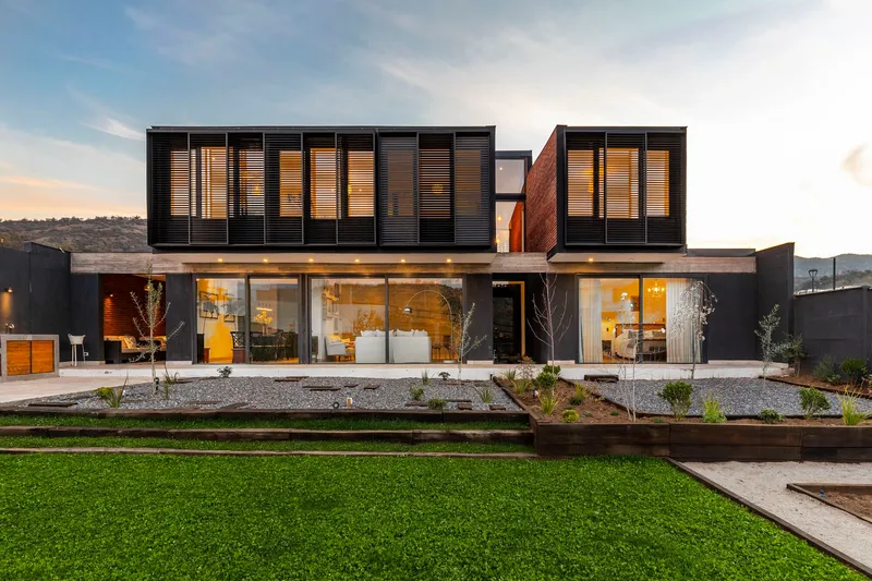 Garden facade with planted beds and floor-to-ceiling glazing beneath slatted black upper volume at twilight