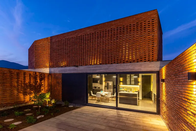 Courtyard view of glazed dining room beneath curved perforated brick volume with planted bed at twilight