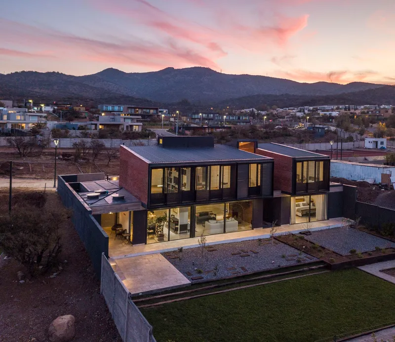 Elevated view of the residence with red brick and glass volumes at dusk with mountains in the background