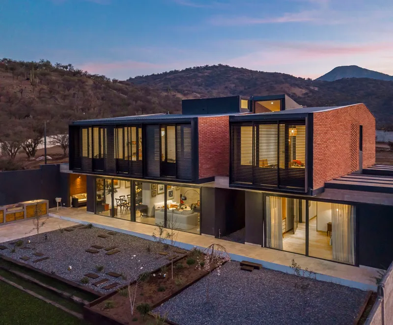 Two-story residence with red brick upper volume and louvered metal facade illuminated at twilight with hillside behind
