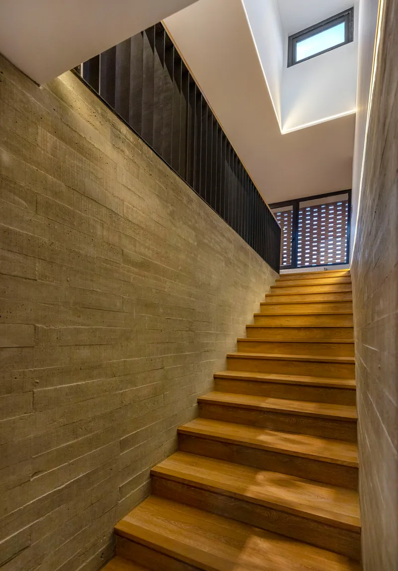 Timber staircase between board-formed concrete walls with skylight and perforated brick screen above