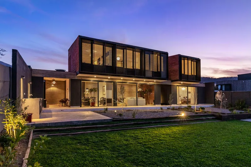 Rear facade at dusk with illuminated glass ground floor and cantilevered brick and metal upper volumes overlooking lawn