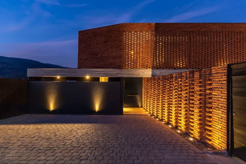 Perforated brick entry wall and canopy lit from below at dusk with paved courtyard