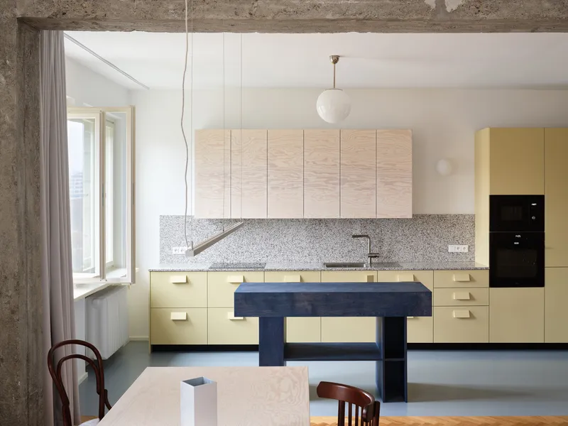 Kitchen with pale yellow cabinets, terrazzo backsplash and a freestanding blue island beneath exposed concrete beams