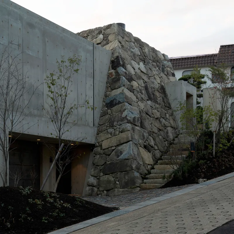 Exposed stone outcrop integrated into a concrete facade with planted beds and young trees below