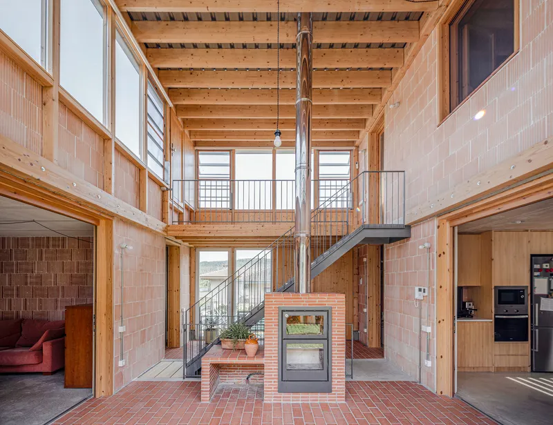 Double-height interior with exposed timber beams and a freestanding brick fireplace beneath a steel flue pipe
