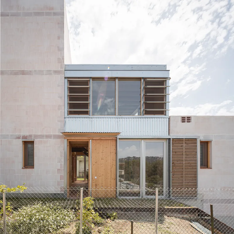 Street facade with corrugated metal cladding, timber panels, and horizontal louvered shutters behind a wire fence