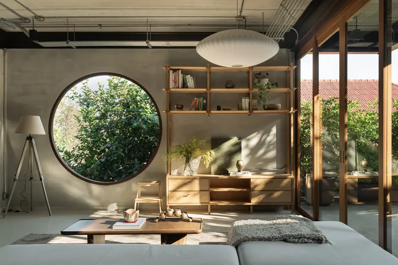 Living room with circular window in concrete wall and timber shelving in afternoon sunlight
