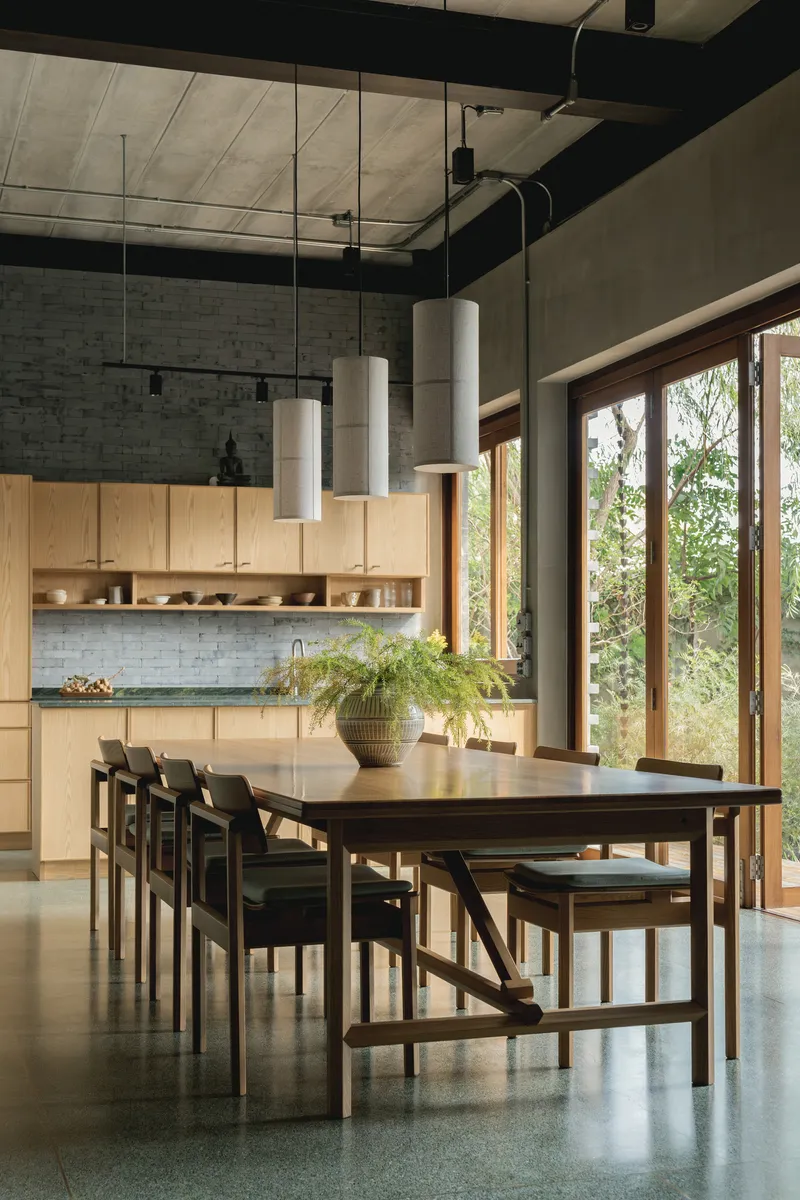 Dining area with exposed concrete ceiling, pendant lights above a timber table, and folding doors to greenery