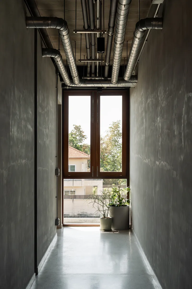 Concrete corridor with exposed ductwork overhead leading to glazed doors and potted plants
