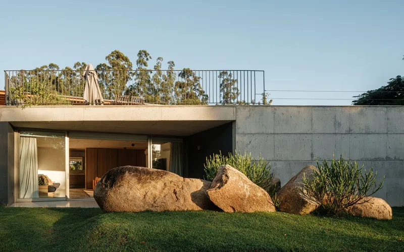 Concrete volume with rooftop garden and large boulders flanking the glass-walled entry in afternoon light