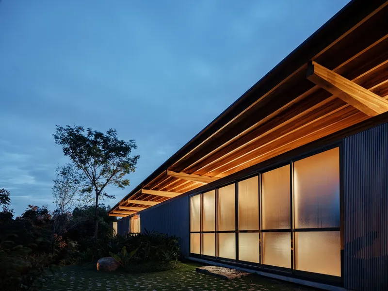 Cantilevered timber roof with exposed beams over corrugated metal facade and translucent panel wall at dusk