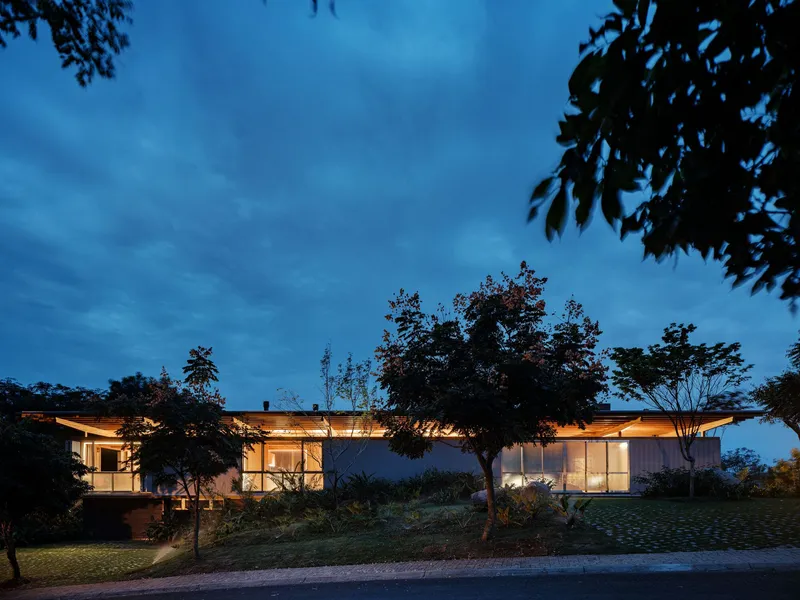 Low-slung residence with illuminated interiors and broad roof overhang seen through mature trees at twilight