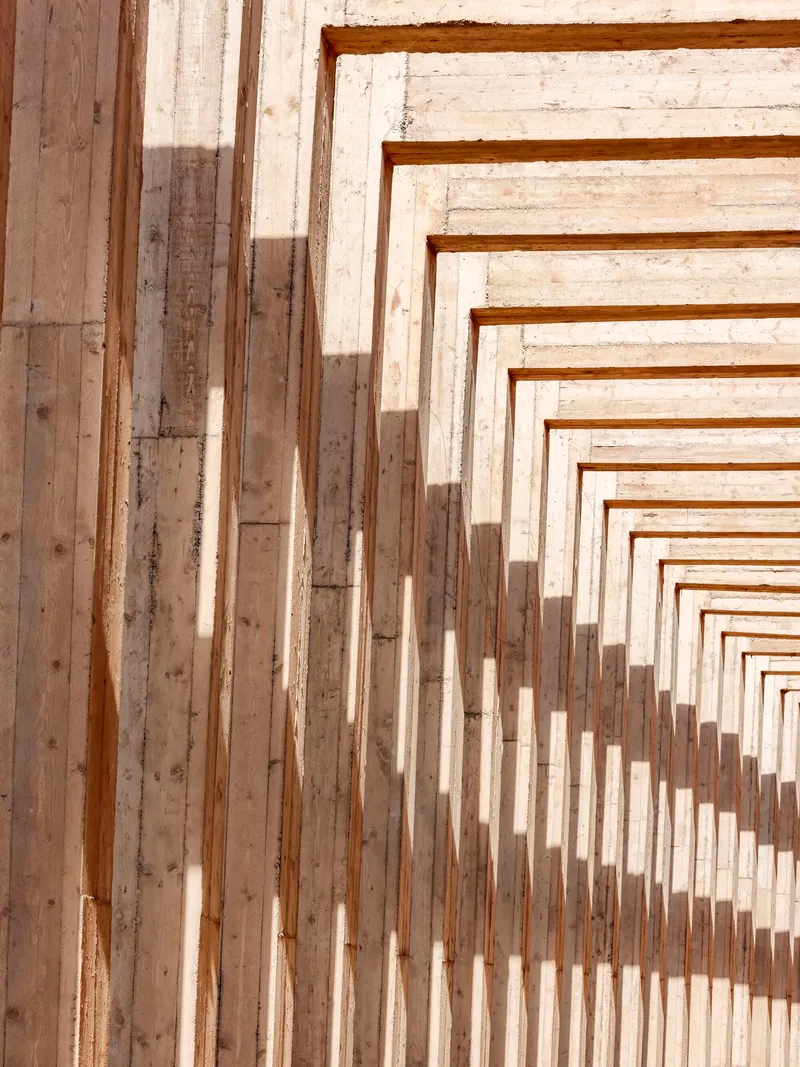 Stepped timber beams forming a rhythmic pergola structure casting shadows in bright sunlight