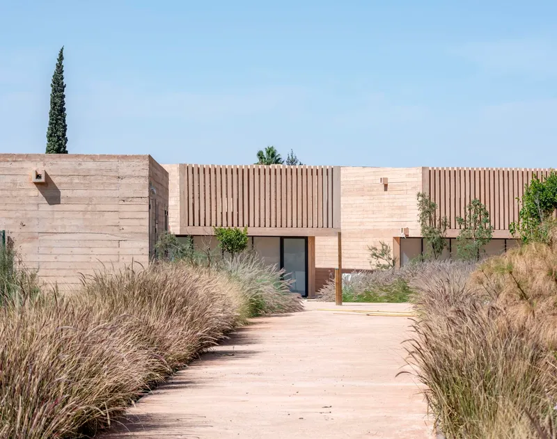 Entrance pathway bordered by wild grasses leading to rammed earth walls and vertical timber screens